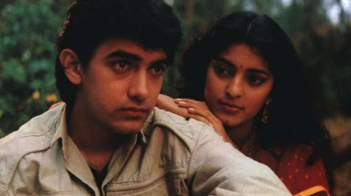 A young Indian woman rests her hands on a young Indian man’s shoulders and gazes lovingly at him as he stares off into the distance